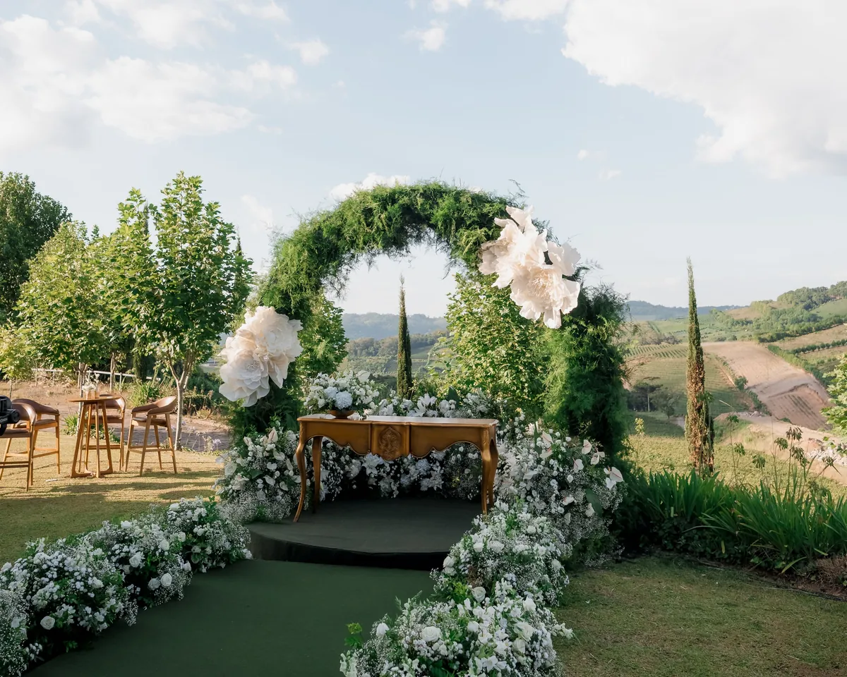 A ceremony set and ready: greenery arch, white florals lining the aisle and a vineyard view