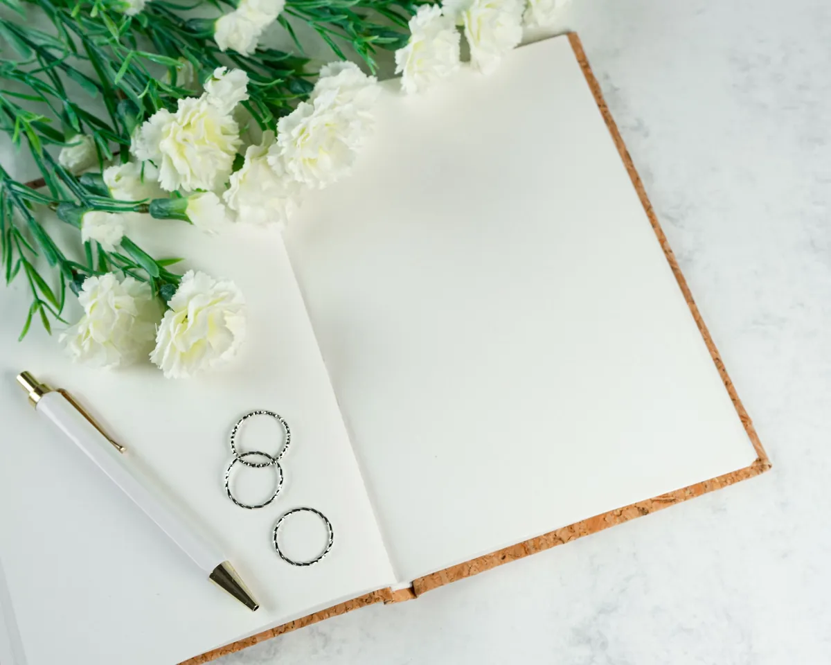 An open planning notebook with wedding rings, a pen and white carnations on marble
